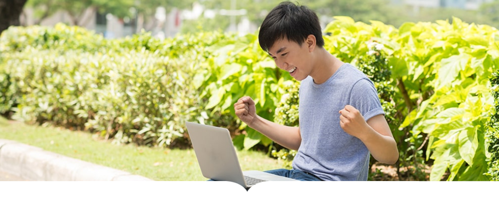 A teenage boy sits in a park on a curb with a laptop on their lap cheering